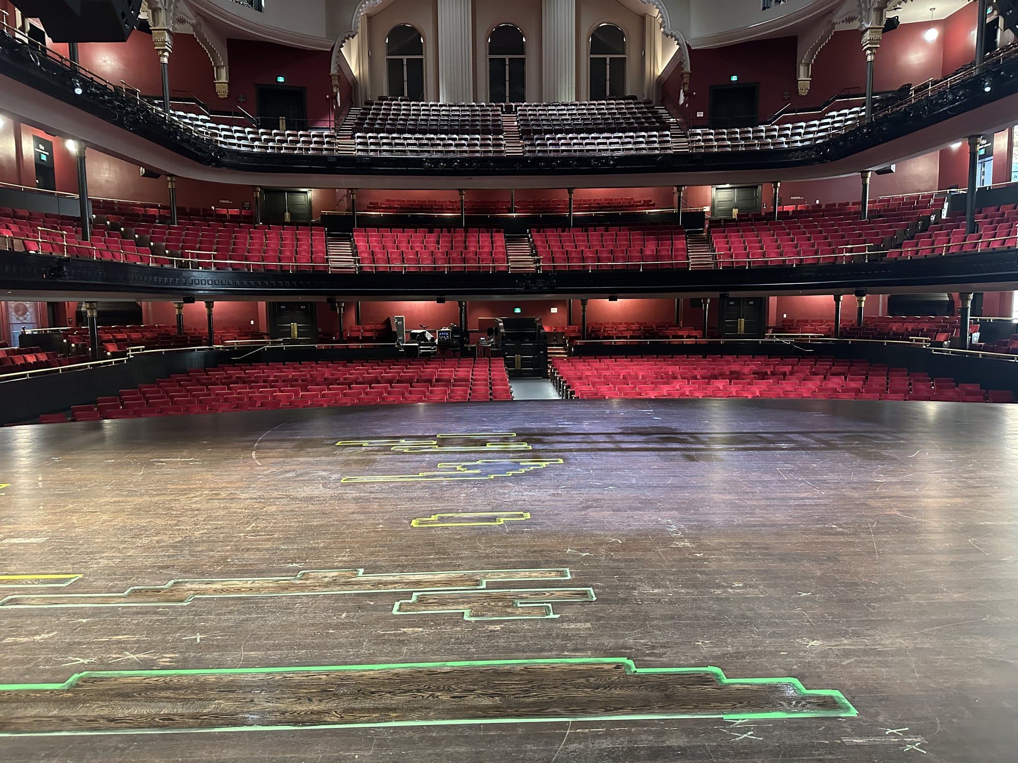 A large wood stage floor in a grand historic theatre, looking out over tiers of red velvet seating